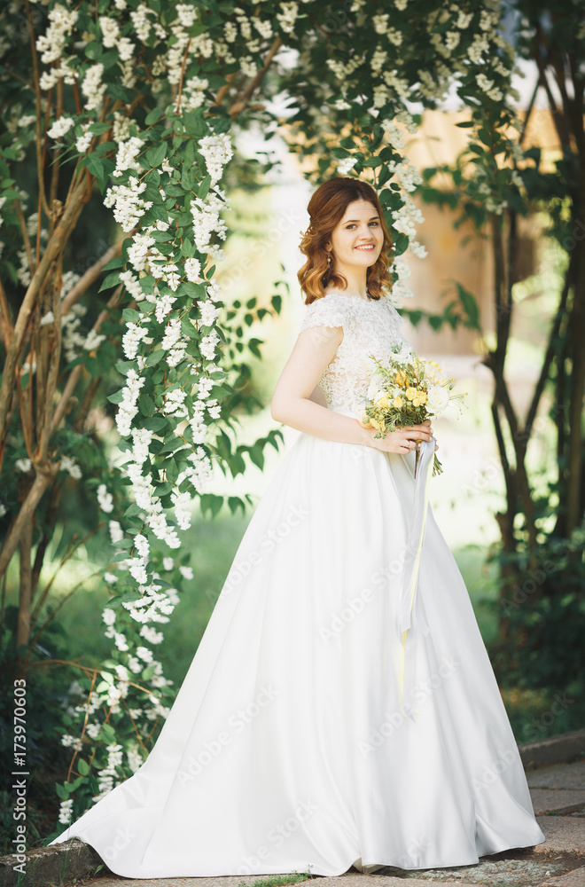 Beautiful brunette bride in elegant white dress holding bouquet posing neat trees