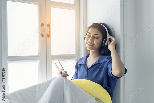 Asian women listen to music in their wireless headphones on the window with a happy smile.