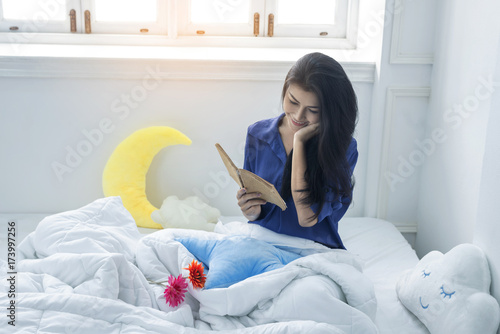 An Asian girl reads a book on her bed in the morning.