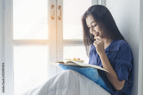 happiness woman enjoy eating chips while reading a book beside window at home in the morning. chill out concept