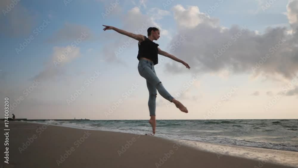 Young woman in casual style - jeans and black top doing ballet at the beach. Attractive ballerina practices in jumping on sandy coastline in autumn. Slow motion. Young woman in casual style - jeans and black top doing ballet at the beach. Attractive ballerina practices in jumping on sandy coastline in autumn. Slow motion.