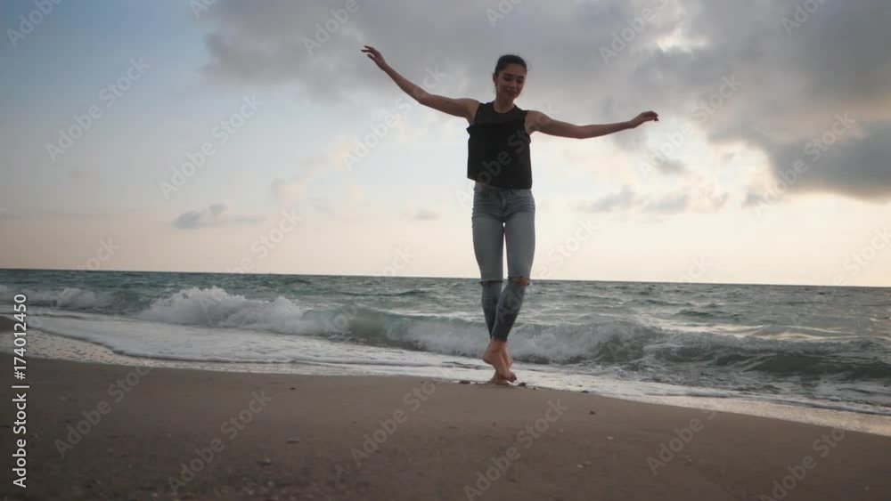 Young woman in casual style - denim and black top doing ballet at the beach. Attractive ballerina practices in stretching on sandy coastline in autumn. Slow motion. Young woman in casual style - denim and black top doing ballet at the beach. Attractive ballerina practices in stretching on sandy coastline in autumn. Slow motion.