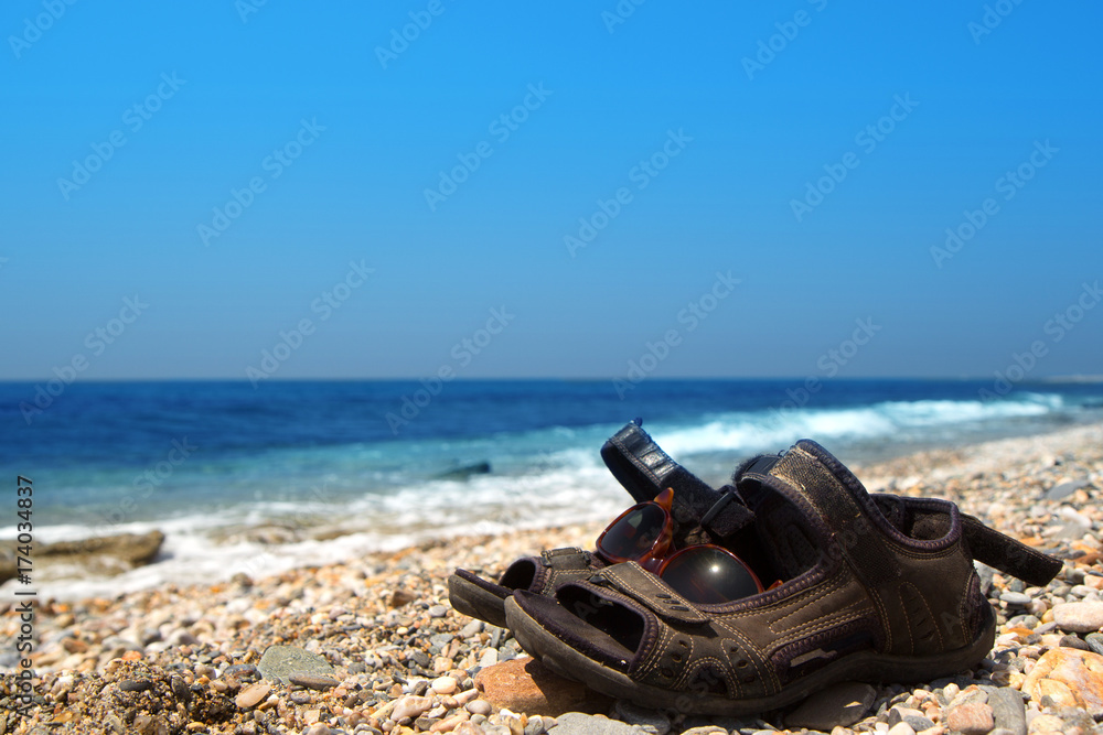 Obraz premium Pair of old sandals and sunglasses on the pebble beach of the Mediterranean Sea