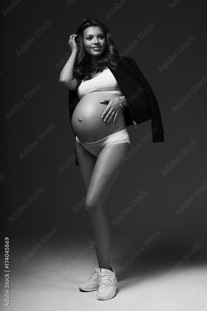 Full body portrait of attractive smiling pregnant brunette woman with slightly curly hair, wearing white sports underwear, trainers and with black jacket draped over shoulders, standing and posing.
