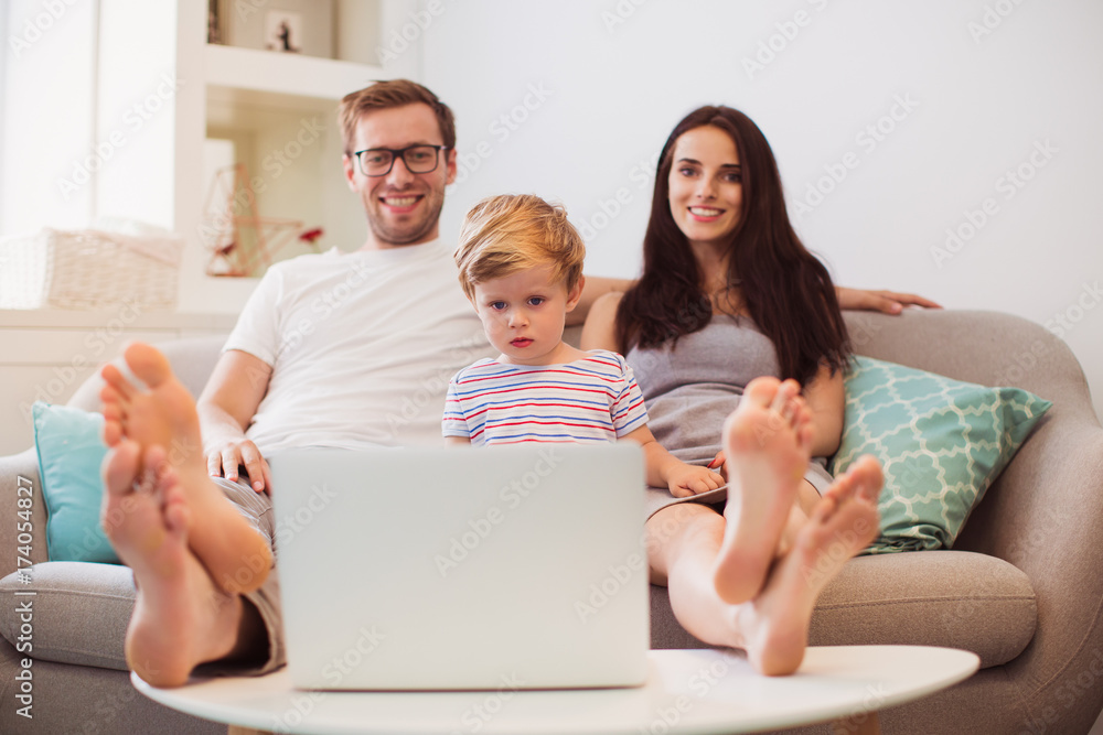 The young happy father, mother and little son sitting on the sofa and watching laptop in the room The young happy father, mother and little son sitting on the sofa and watching laptop in the room
