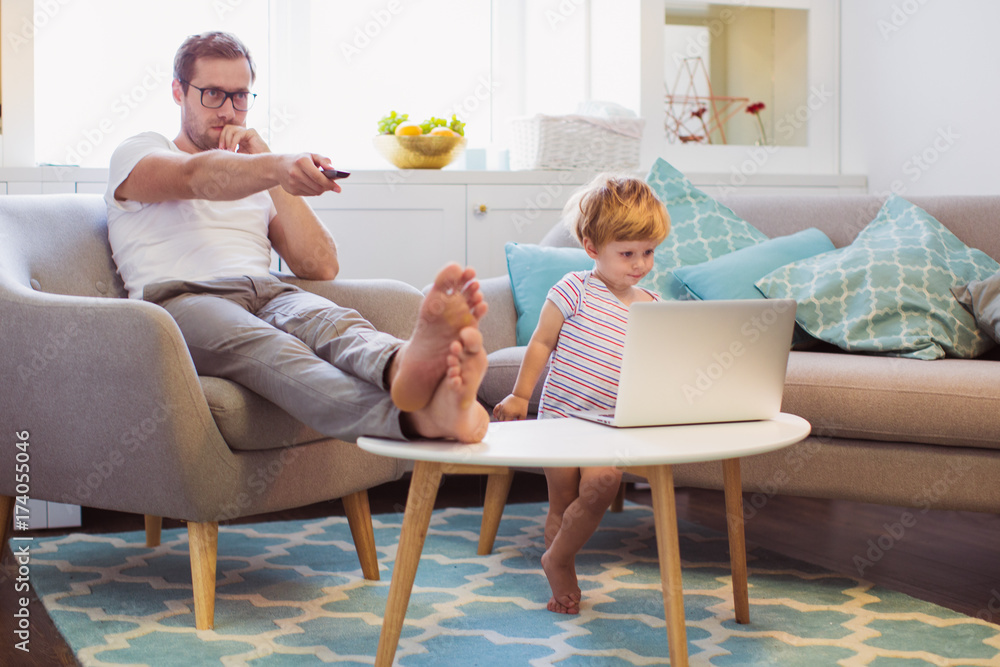 The young serious father sitting on the sofa in the room and turning on TV near his cute little son who watching laptop The young serious father sitting on the sofa in the room and turning on TV near his cute little son who watching laptop