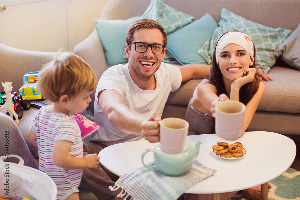 The young smiling parents sitting at the table in the room and keeping cups of tea near little son who playing The young smiling parents sitting at the table in the room and keeping cups of tea near little son who playing
