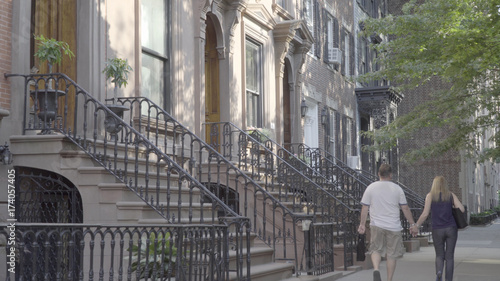 DX exterior establishing shot of a typical generic Brooklyn brownstone home row. Famous steps lead to front door of expensive real estate. Couple on romantic walk sidewalk past houses