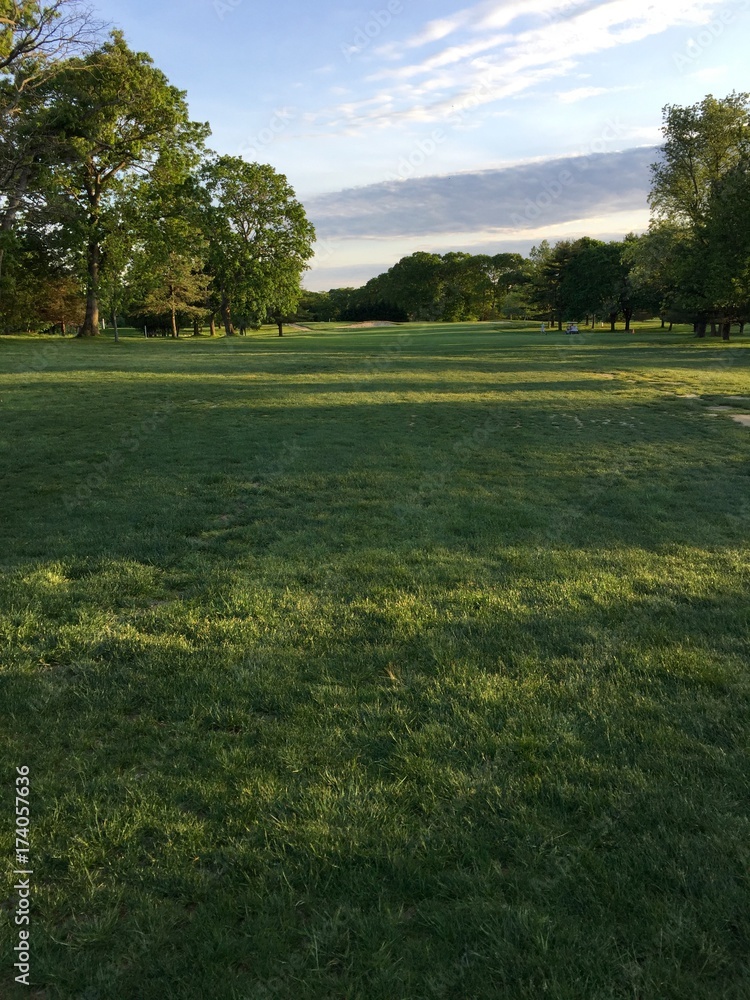 View down the fairway of a beautiful green generic golf course at sunset. Sun light peaks between trees to cast shadows across landscape. Vertical photo.