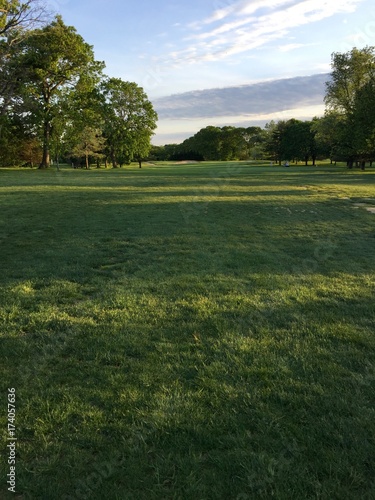 Obraz na płótnie View down the fairway of a beautiful green generic golf course at sunset. Sun light peaks between trees to cast shadows across landscape. Vertical photo. w salonie
