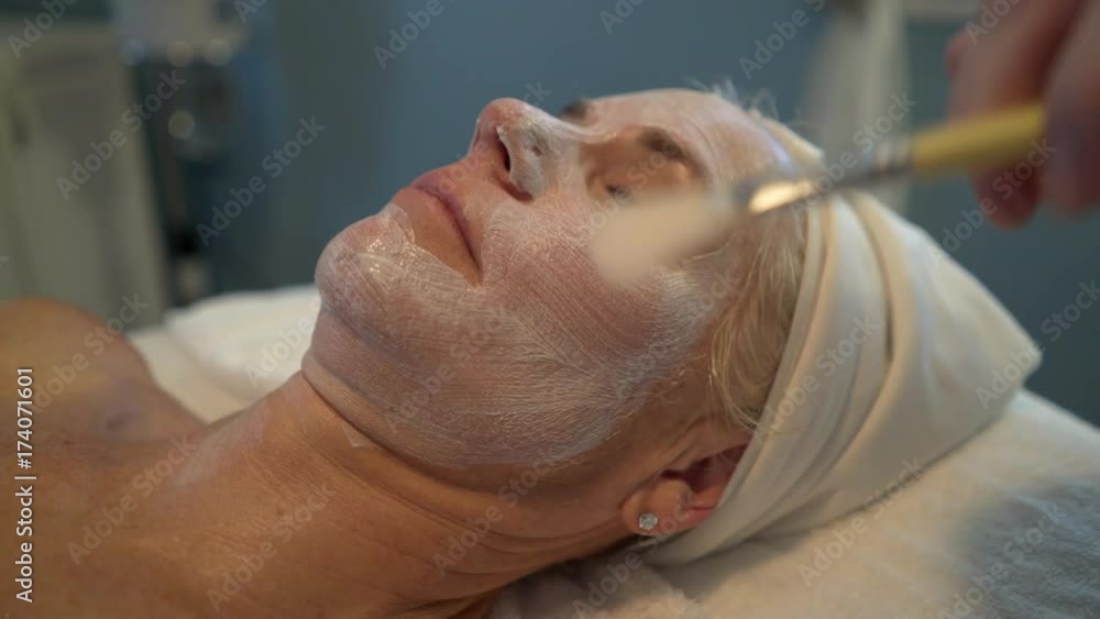 Massage therapist brushes a cream onto a beautiful mature woman’s face as part of a facial. Steadicam shot. Massage therapist brushes a cream onto a beautiful mature woman’s face as part of a facial. Steadicam shot.