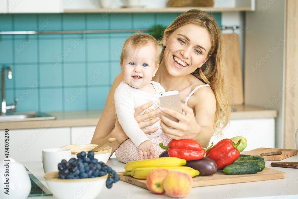 Young beautiful mother is cooking and playing with her baby daughter in a modern kitchen setting. Healthy food concept. Taking selfie on camera.