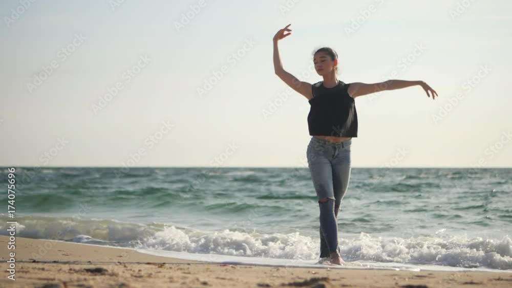 Young woman in casual style - denim and black top doing ballet at the beach. Attractive ballerina practices in hands exercises on sandy plage in autumn. Slow motion. Sea waves background Young woman in casual style - denim and black top doing ballet at the beach. Attractive ballerina practices in hands exercises on sandy plage in autumn. Slow motion. Sea waves background