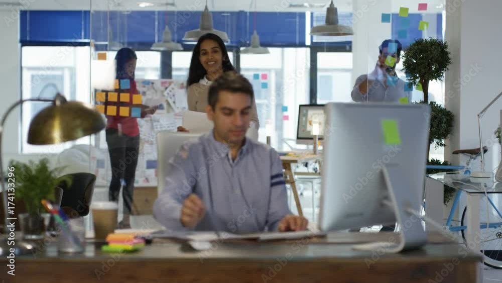 Designer working at his desk in creative office and talking to female colleague. Designer working at his desk in creative office and talking to female colleague.