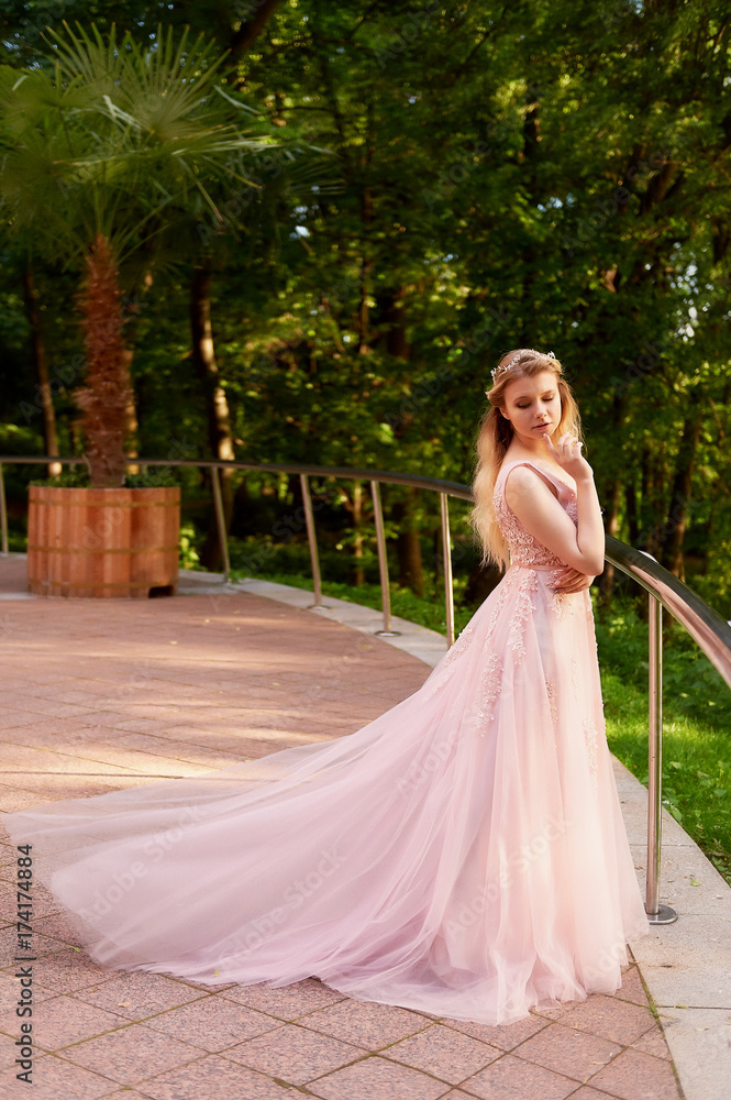Portrait of a beautiful bride blond girl in pink lace dress ,hair decoration, handmade. Tenderness. Standing in the midst of a dark forest green in the rays of the setting sun. Shows a long dress with