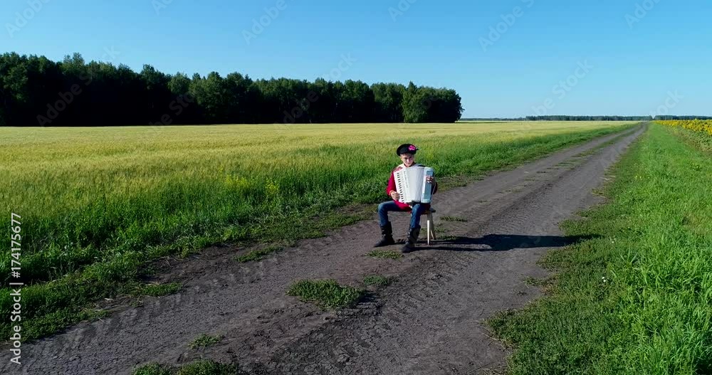 Aerial: A young man in a Russian traditional costume plays the accordion sitting on a chair on a country road, between a wheat and sunflowers field. Forest on the background. Aerial: A young man in a Russian traditional costume plays the accordion sitting on a chair on a country road, between a wheat and sunflowers field. Forest on the background.