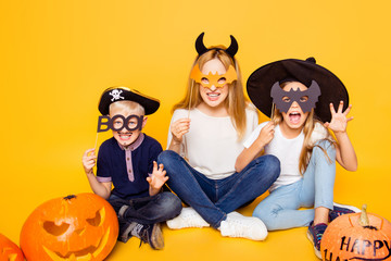 Close up portrait of mom with two small monsters, hiding behind eyemasks, with frightening gestures, sitting on the floor with crossed legs, handmade cutted pumpkin near, isolated on background