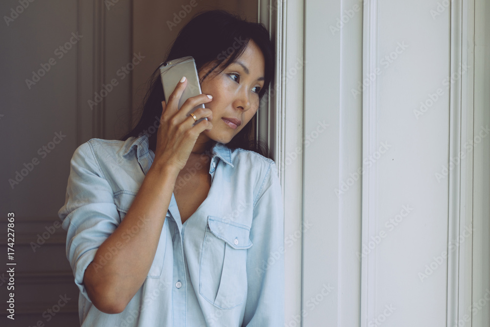 Beautiful asian woman with long dark hair is talking by a smartphone while looking thoughtfully out the window. Hipster girl is chatting with friends by a mobile phone while standing beside the window Beautiful asian woman with long dark hair is talking by a smartphone while looking thoughtfully out the window. Hipster girl is chatting with friends by a mobile phone while standing beside the window