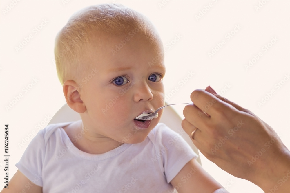Mother feedding her one year blonde blue-eyed toddler with a yogurt Mother feedding her one year blonde blue-eyed toddler with a yogurt