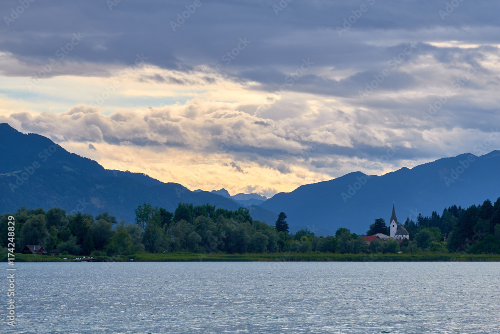 A church with white tower on the shore of Faaker See, in Austria, in front of beautiful sunset sky