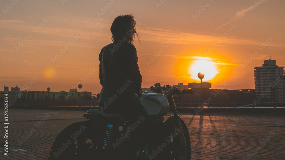 Fototapeta premium Outdoor lifestyle portrait of sexy biker girl wearing leather jacket sits on a modern motorcycle, sunset over city in the background