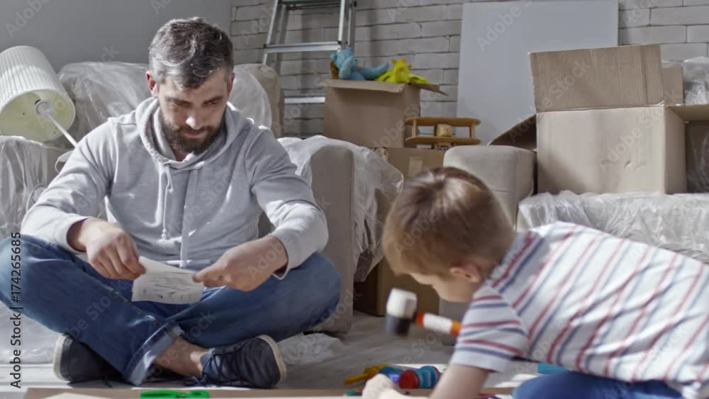Zoom out shot of little boy playing with toy hummer and disassembled furniture while bearded man sitting and reading manual Zoom out shot of little boy playing with toy hummer and disassembled furniture while bearded man sitting and reading manual