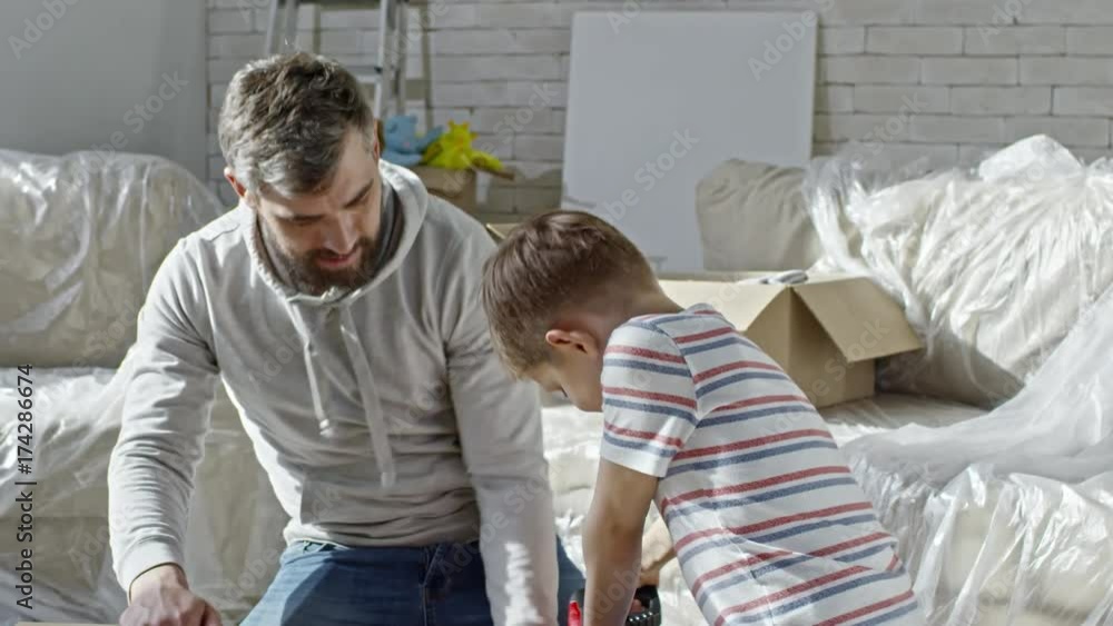Zoom out shot of bearded father sitting on floor of new apartment and showing little son how to put screws in with electric screwdriver Zoom out shot of bearded father sitting on floor of new apartment and showing little son how to put screws in with electric screwdriver