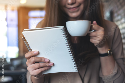 Closeup image of a beautiful Asian woman with smiley face holding and looking at a white blank notebook while drinking coffee in modern cafe