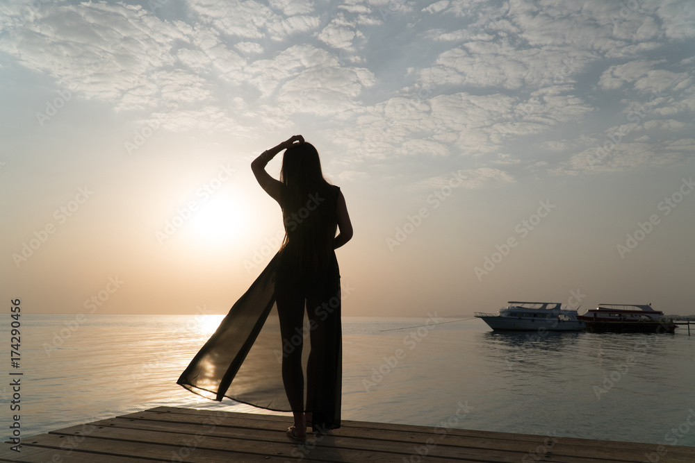 A girl in a swimsuit and a cloak is standing on the pier and meets the dawn