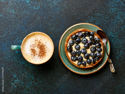 Blueberry tart with cup of coffee on blue stone background.