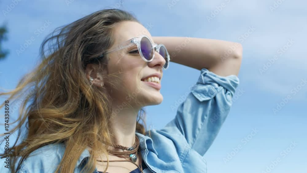 Stylish Young Girl with Brown Hair Wearing Cool Sunglasses Smiles Charmingly on Camera. She Runs Hand Through Her Beautiful Long Hair. Clear Blue Sky visible in the Background. Stylish Young Girl with Brown Hair Wearing Cool Sunglasses Smiles Charmingly on Camera. She Runs Hand Through Her Beautiful Long Hair. Clear Blue Sky visible in the Background.