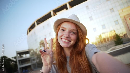 charming ginger girl in hat taking selfie video having fun fooling around showing peace with stadium background slomo closeup