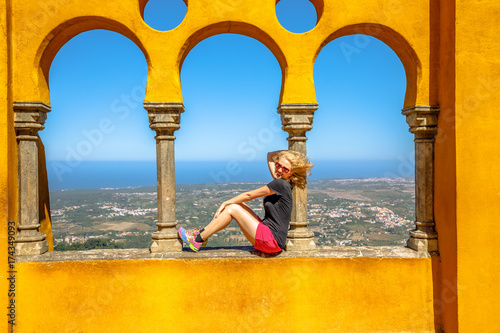 Happy blonde woman sitting on arabic arches of Pena National Palace on top of a hill above Sintra.On background, aerial view of the city.Female traveler visits the biggest tourist attraction of Sintra photo