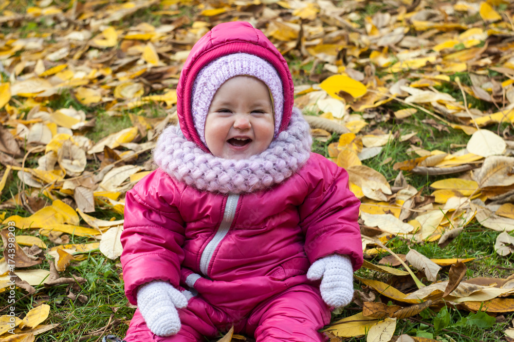 smiling girl in pink jumpsuit, white mittens and pink scarf sitting in the autumn on the nature walk outdoors smiling girl in pink jumpsuit, white mittens and pink scarf sitting in the autumn on the nature walk outdoors
