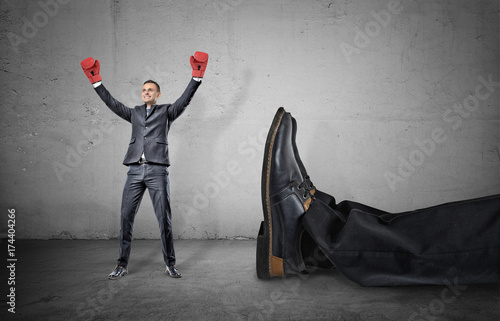 A happy businessman with boxing gloves on arms raised in victory stands near a giant male leg fallen down.