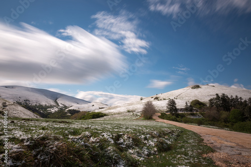 Long exposure photo of a mountain scenery with green grass and melting snow near a road, under a blue sky with moving clouds