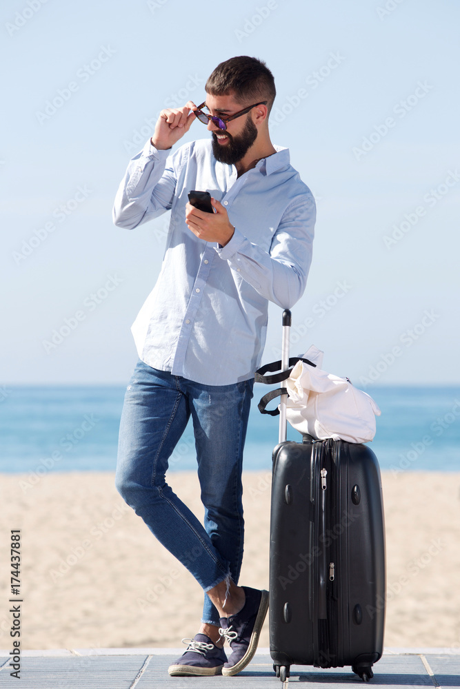 Obraz premium Full length man standing on beach with suitcase and mobile phone
