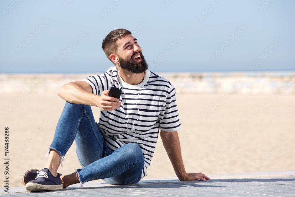 Full length happy man in earphones sitting on beach with mobile phone