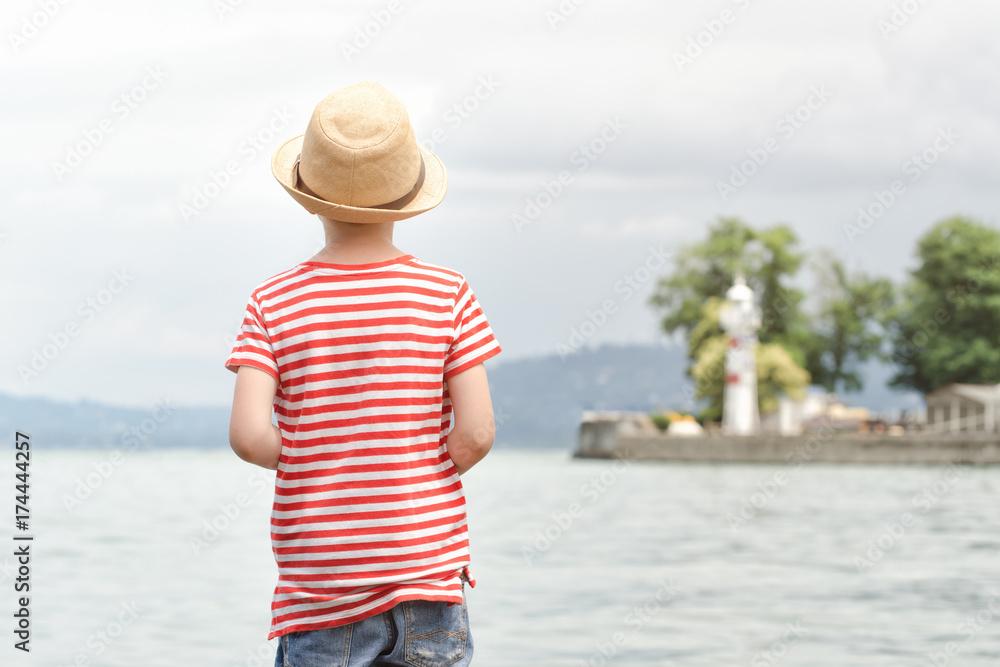 Boy in a hat and striped t-shirt standing on the beach. View from the back