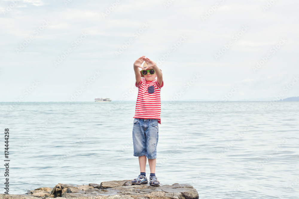 Boy in a hat and a striped T-shirt standing on a rock by the sea