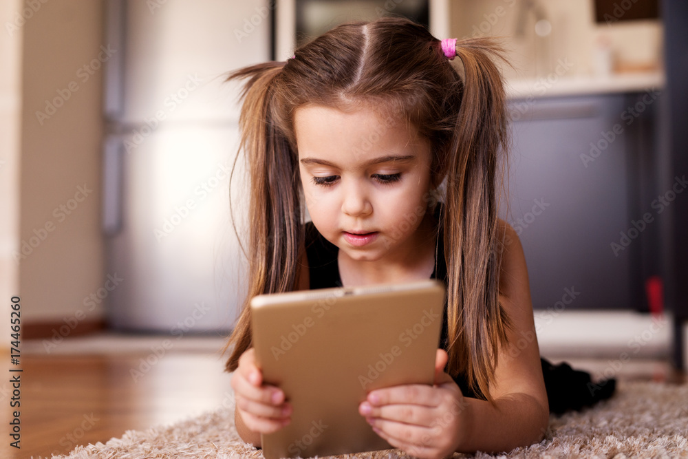 Beautiful child little girl with ponytails playing with a tablet while lying on the floor. Beautiful child little girl with ponytails playing with a tablet while lying on the floor.