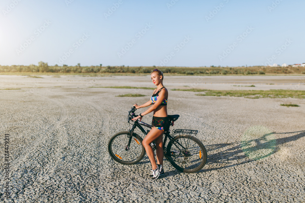 A strong blonde woman in a multicolored suit stands near a bicycle in a desert area and looks at the sun. Fitness concept. Blue sky background