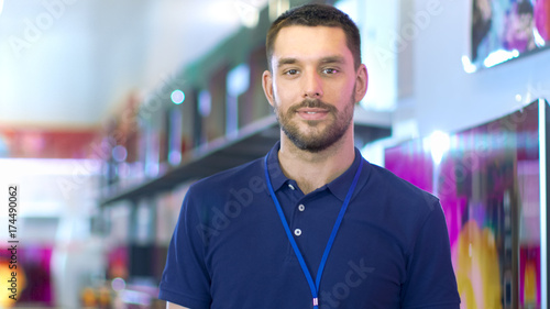 Portrait of a Professional Expert Consultant Smiles and Looks into Camera as Stands in the Bright, Modern Electronics Store Full of Latest Models of TV Sets, Cameras, Tablets and other Devices. photo