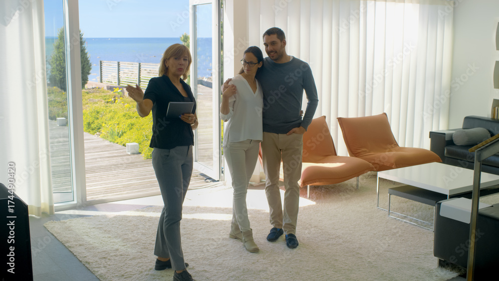 Professional Real Estate Agent Shows Stylish Modern House to a Beautiful Young Couple Who are in the Market for Purchasing/ Renting New Home. House Has Floor to Ceiling Windows and Seaside View.