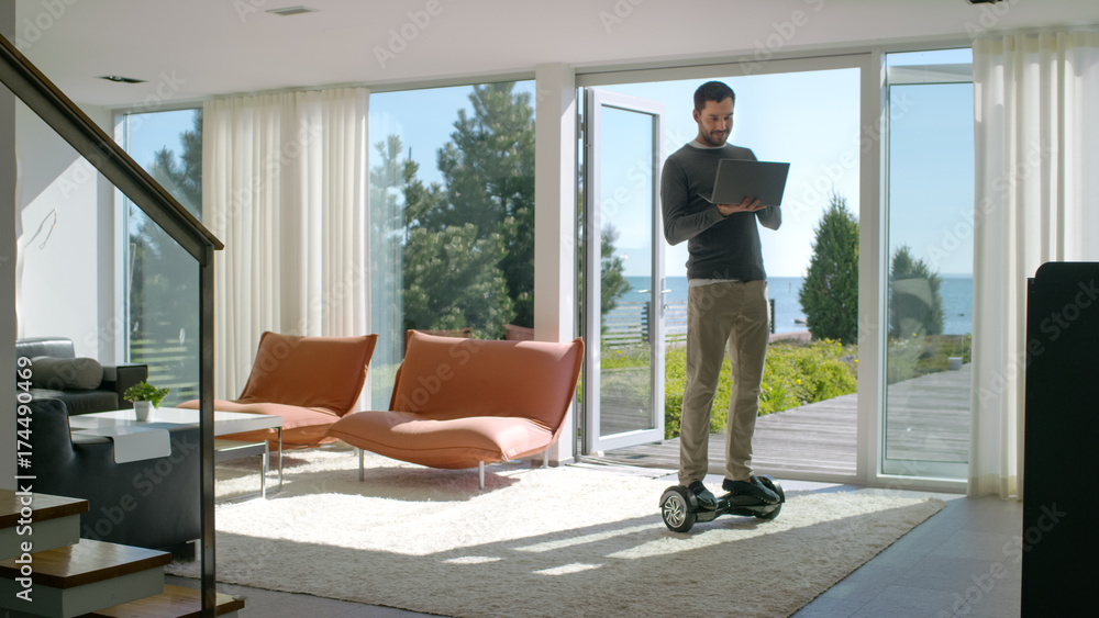 Tech Industry Businessman Working From Home Uses Laptop Computer and Rides Around the House on His Gyro Scooter. His Modern House has Seaside View. Tech Industry Businessman Working From Home Uses Laptop Computer and Rides Around the House on His Gyro Scooter. His Modern House has Seaside View.