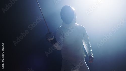 Fully Equipped Fencer Puts Lifts Foil Sword in Readiness for a Match. He Stands in the Spotlight while Darkness is Around Him. Shot Isolated on Black Background.