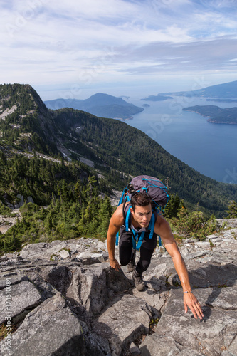 Fit and Adventurous Latin American man is hiking on top of a mountain ridge with a beautiful ocean view in the background. Taken on Lions Peaks, North of Vancouver, British Columbia, Canada. 