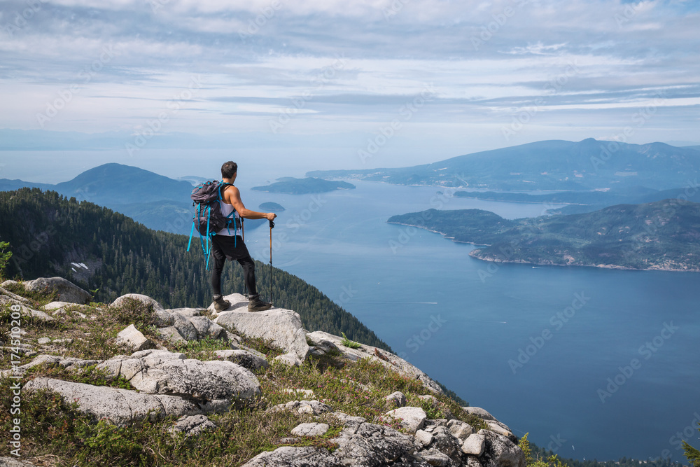 Obraz premium Fit and Adventurous Latin American man is hiking on top of a mountain ridge with a beautiful ocean view in the background. Taken on Lions Peaks, North of Vancouver, British Columbia, Canada. 