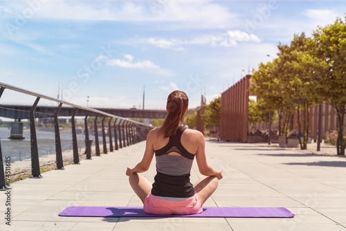 Young woman in lotus position doing yoga in the city in sunny day by the river.