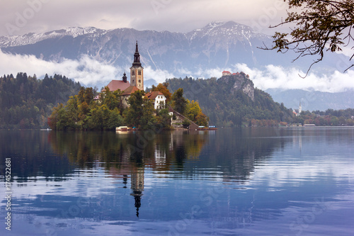 Autumn view of the historical church on the island in Lake Bled in front of snow capped mountains under clouds